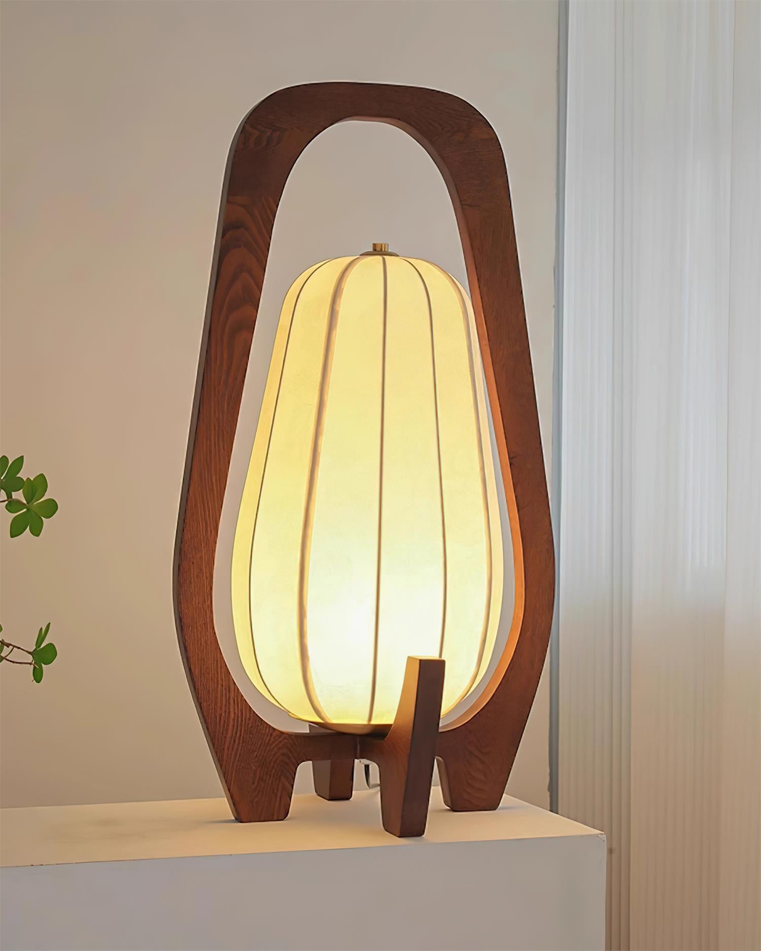 Wooden table lamp with visible natural grain and warm glowing light, eye-level front view with white table and greenery