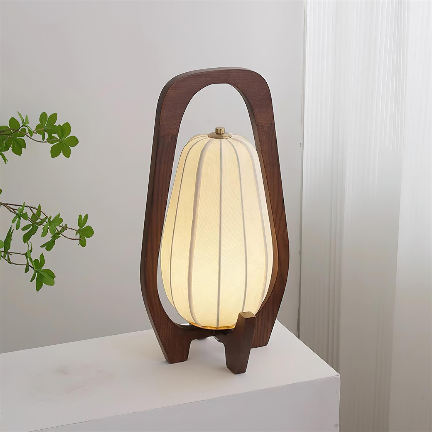 Nordic wooden table lamp with dual-leg wooden base and lit beige fabric shade beside sheer curtains and green plant