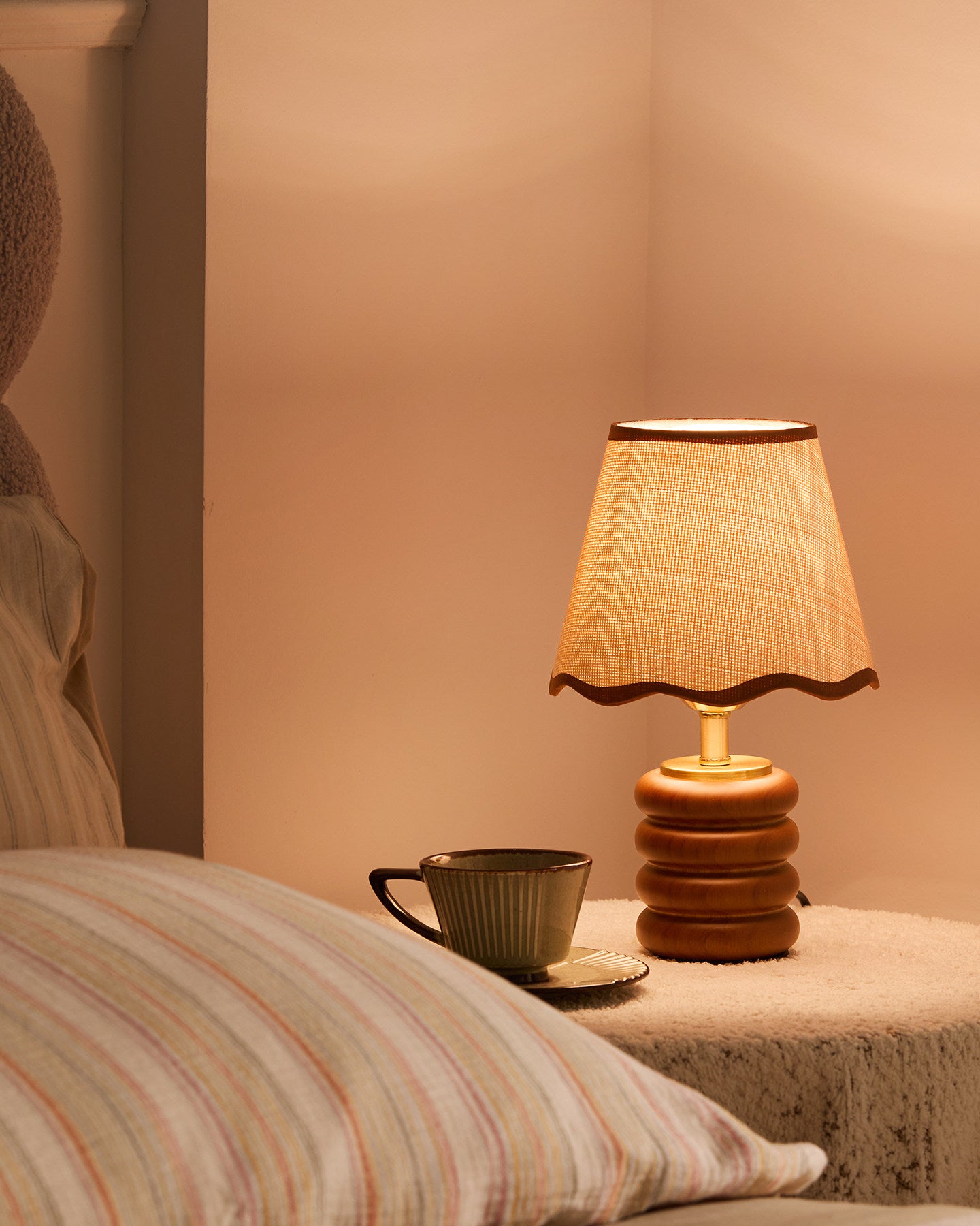 Glowing Marlowe fabric shade table lamp on bedroom side table next to coffee cup and pillow, cozy nighttime lighting