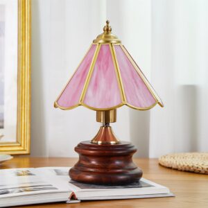 Pink Tiffany style stained glass table lamp on wooden desk with book and curtain backdrop, three-quarter view showing brass hardware