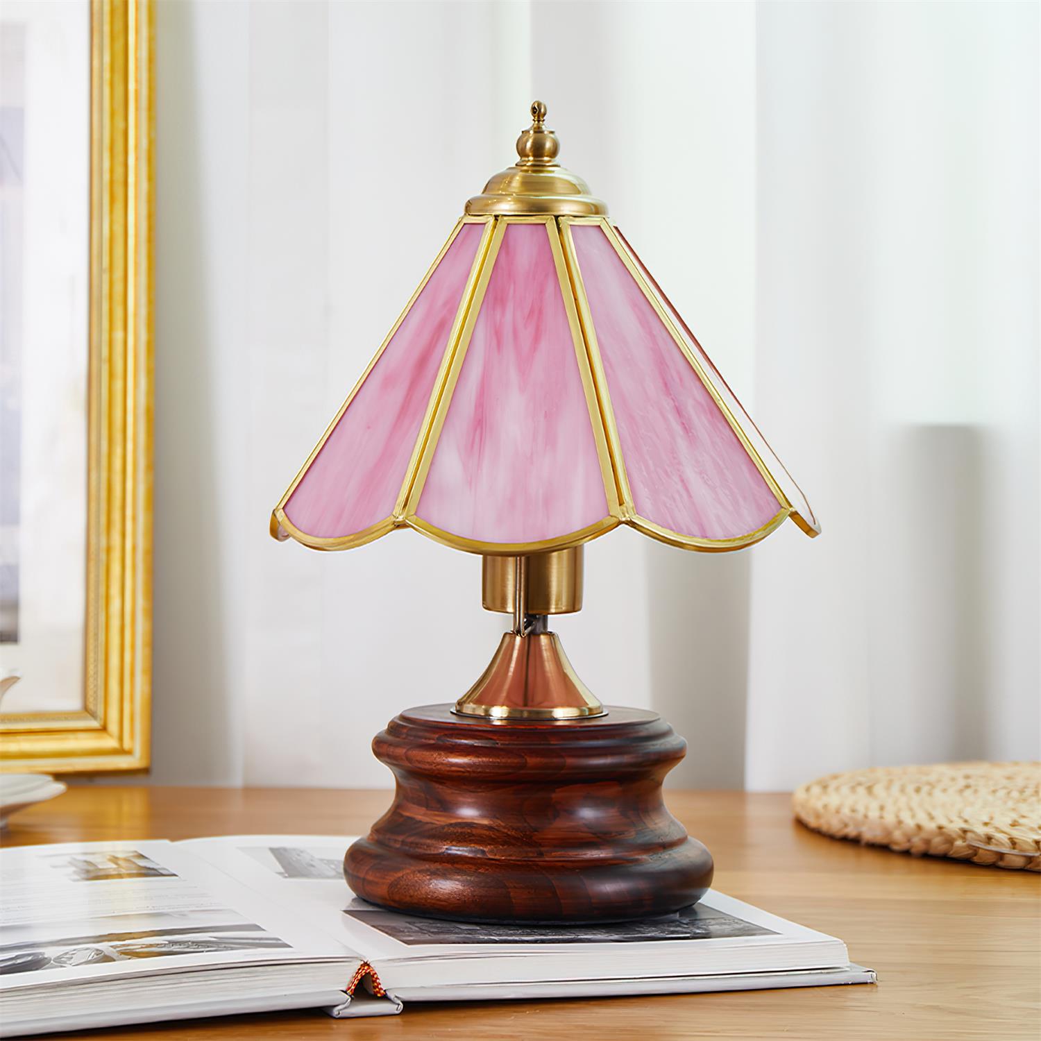 Pink Tiffany style stained glass table lamp on wooden desk with book and curtain backdrop, three-quarter view showing brass hardware
