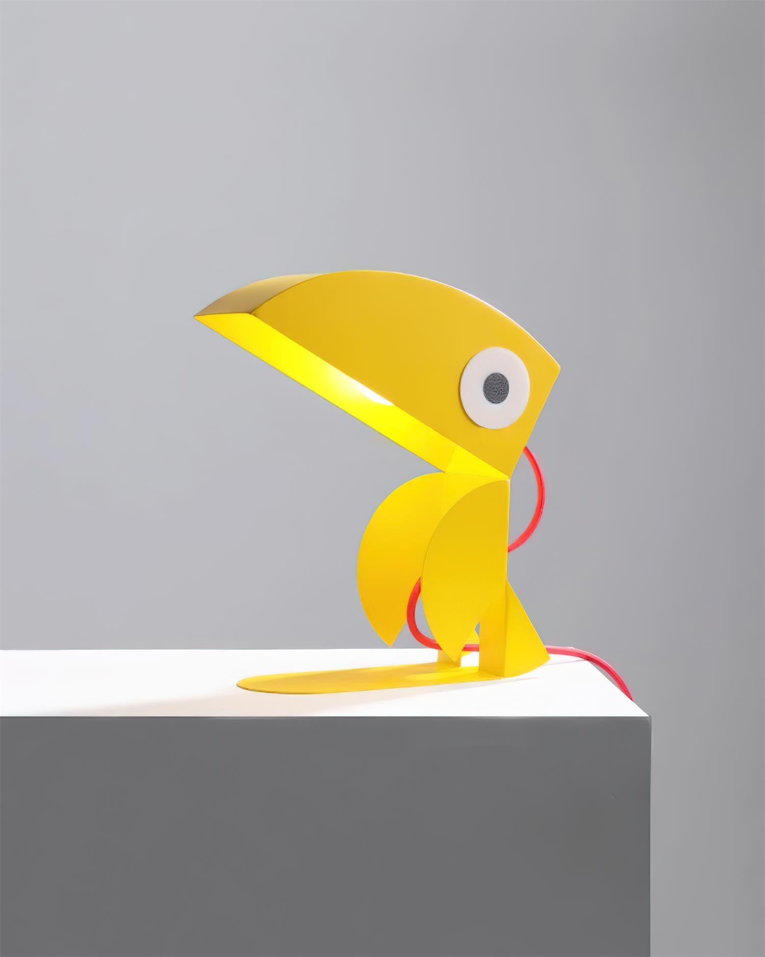 Yellow toucan playful metal table lamp on grey display plinth with warm illumination