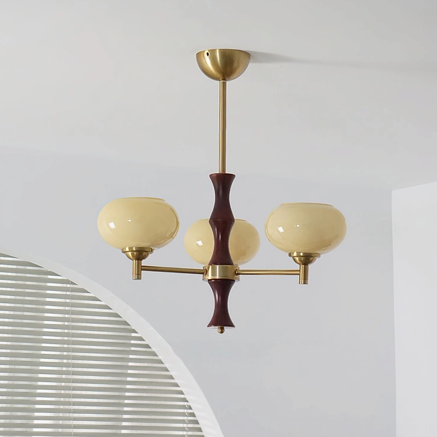 3-head mid-century chandelier with warm beige glass shades, brass arms, and dark wood stem installed in home space with arched louvered wall