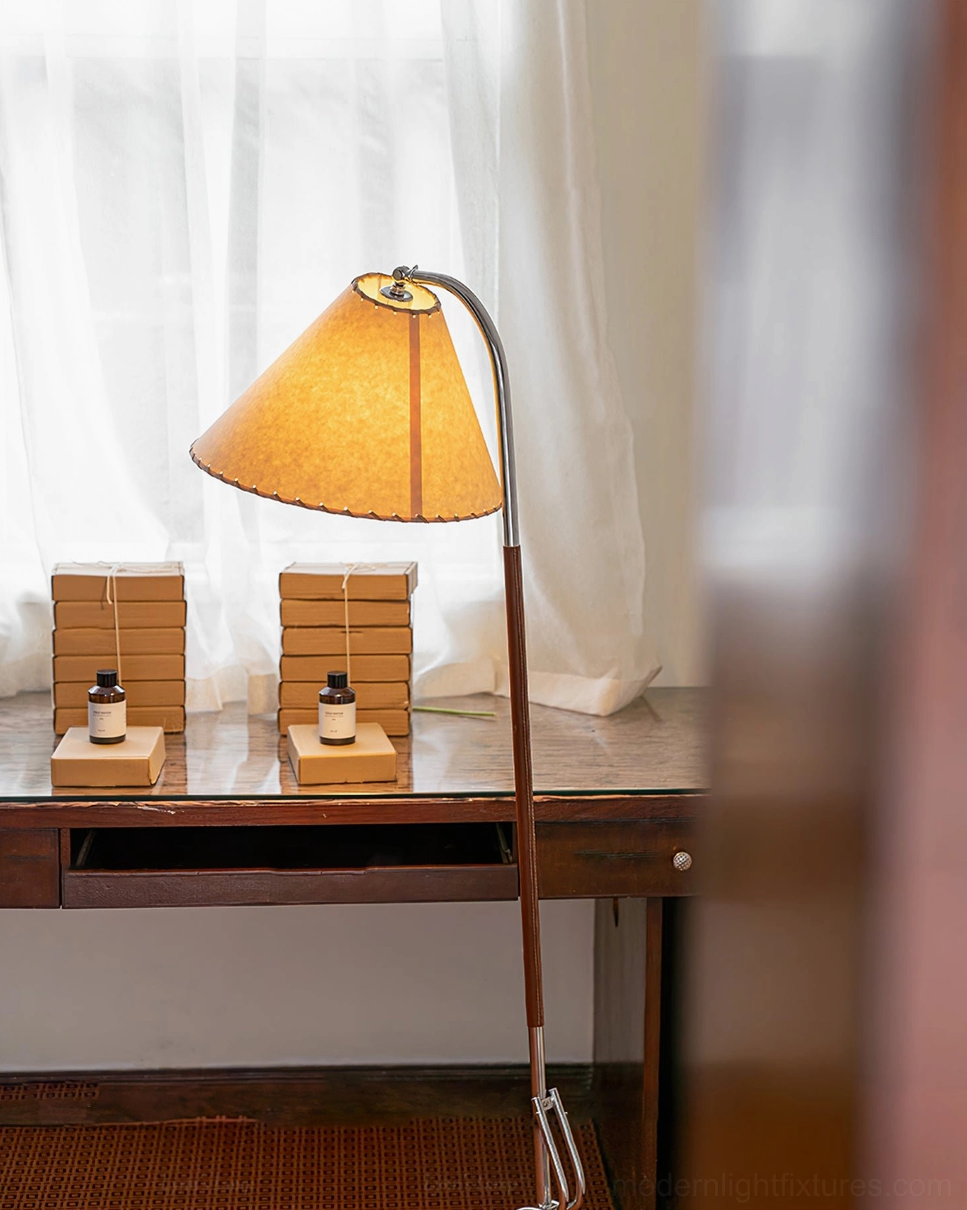 Adjustable tripod floor lamp beside home office desk with sheer window curtains, showing fabric conical shade with stitched trim details and chrome curved arm