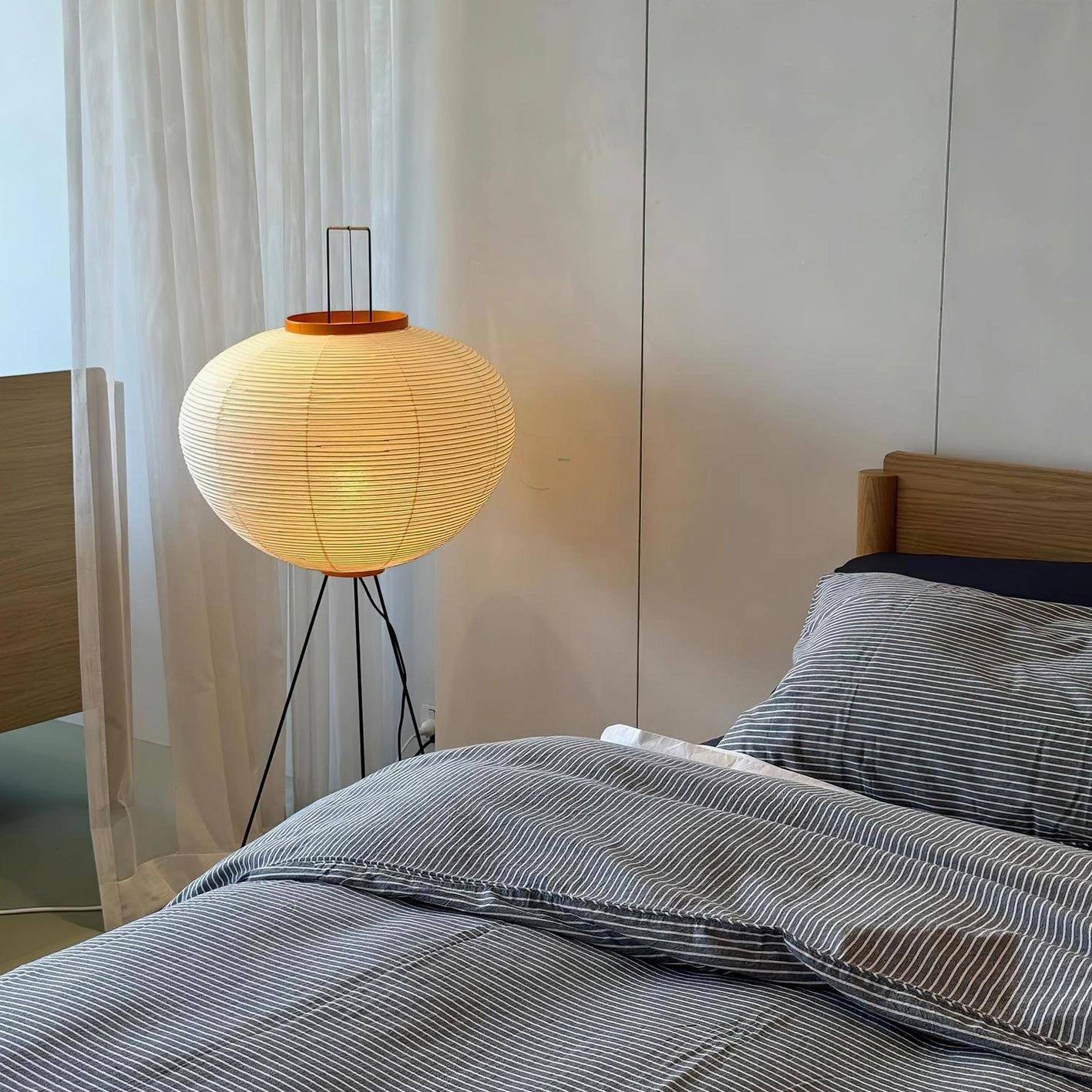 Lit Akari 10A floor lamp in bedroom with warm yellow glow, striped linen bedding and sheer curtains