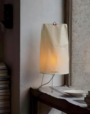 Akari 1P tripod paper lamp placed on bedroom side table by window with warm light glow and stacked books nearby