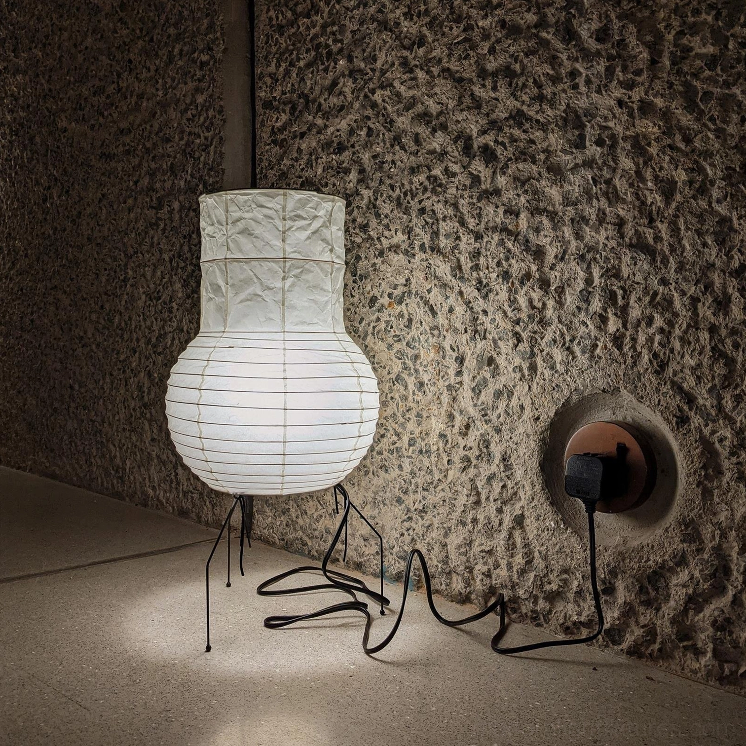 Akari UF1-S white paper table lamp plugged into wall socket, side angle view in corner setting with textured concrete backdrop