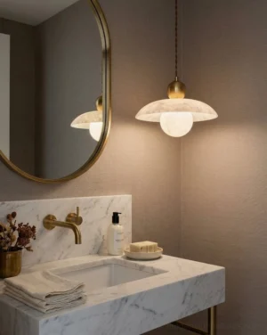 Alabaster pendant light hanging above bathroom vanity with round mirror, warm brass fixtures, and neutral tile backdrop