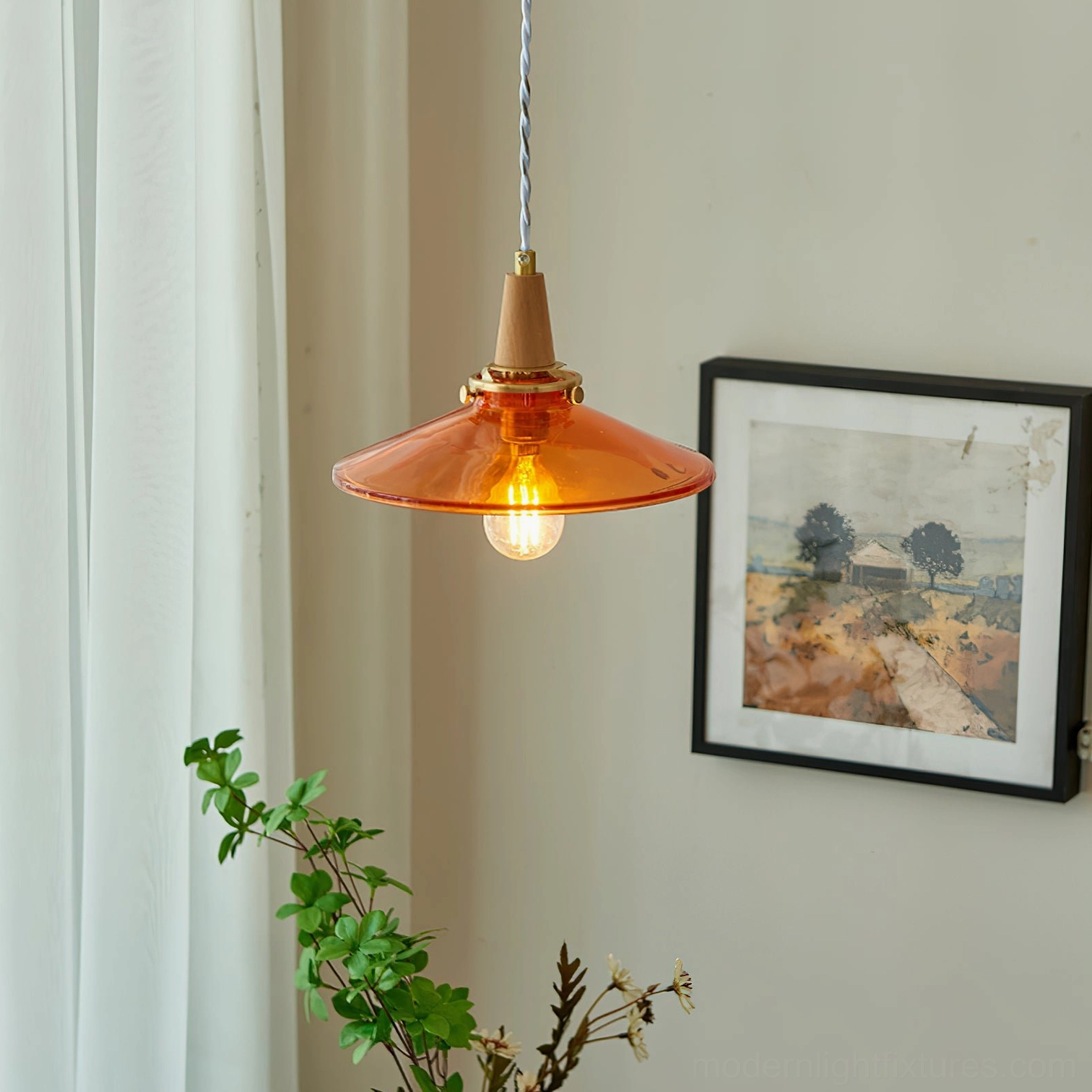 Amber glass pendant light with twisted cloth cord installed in interior space with wall art and green foliage