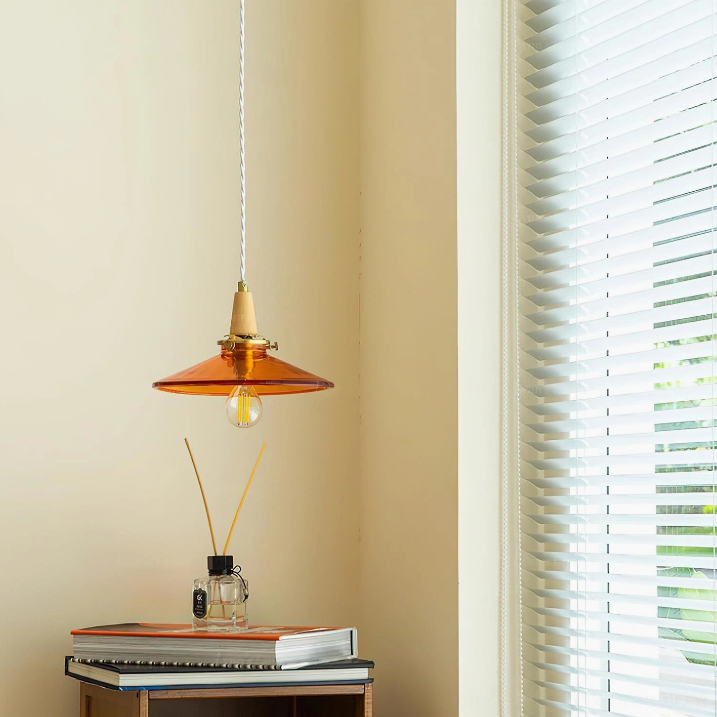 Amber glass pendant lamp side view near louvered window with books and reed diffuser on table
