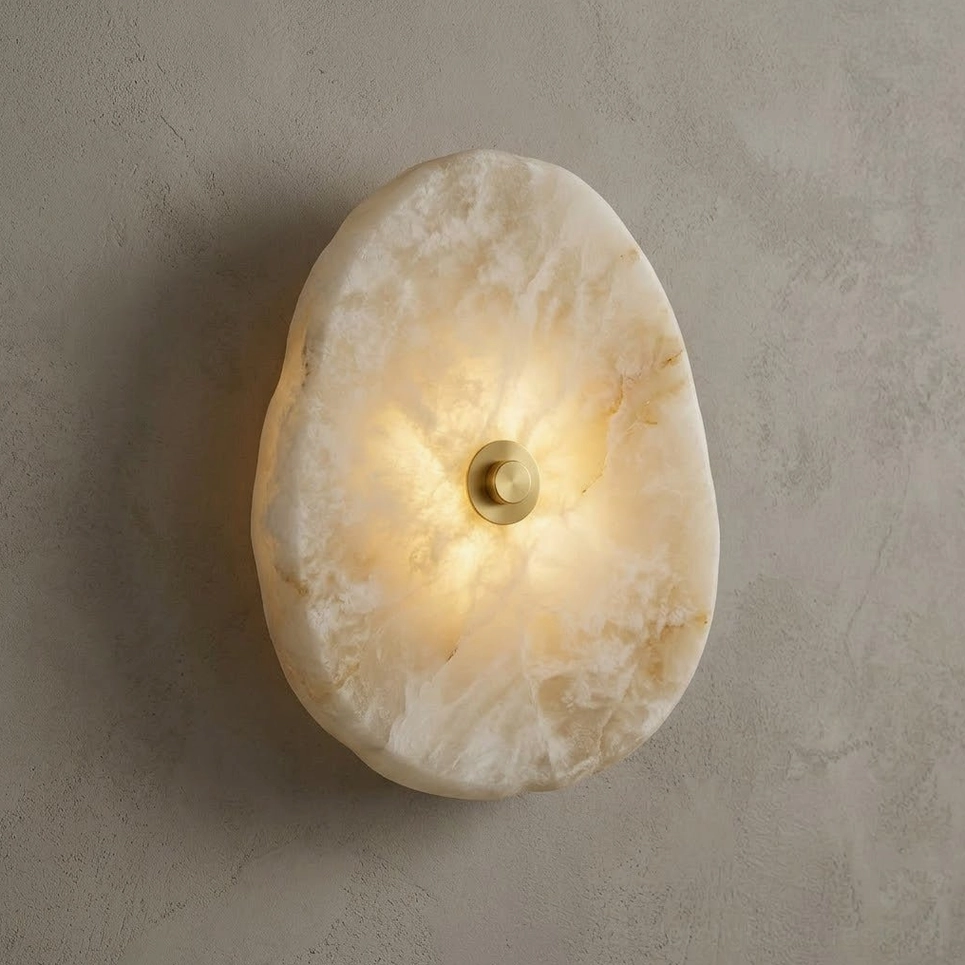 Alabaster wall sconce with brushed brass disc, three-quarter side view on textured concrete wall with warm ambient LED light