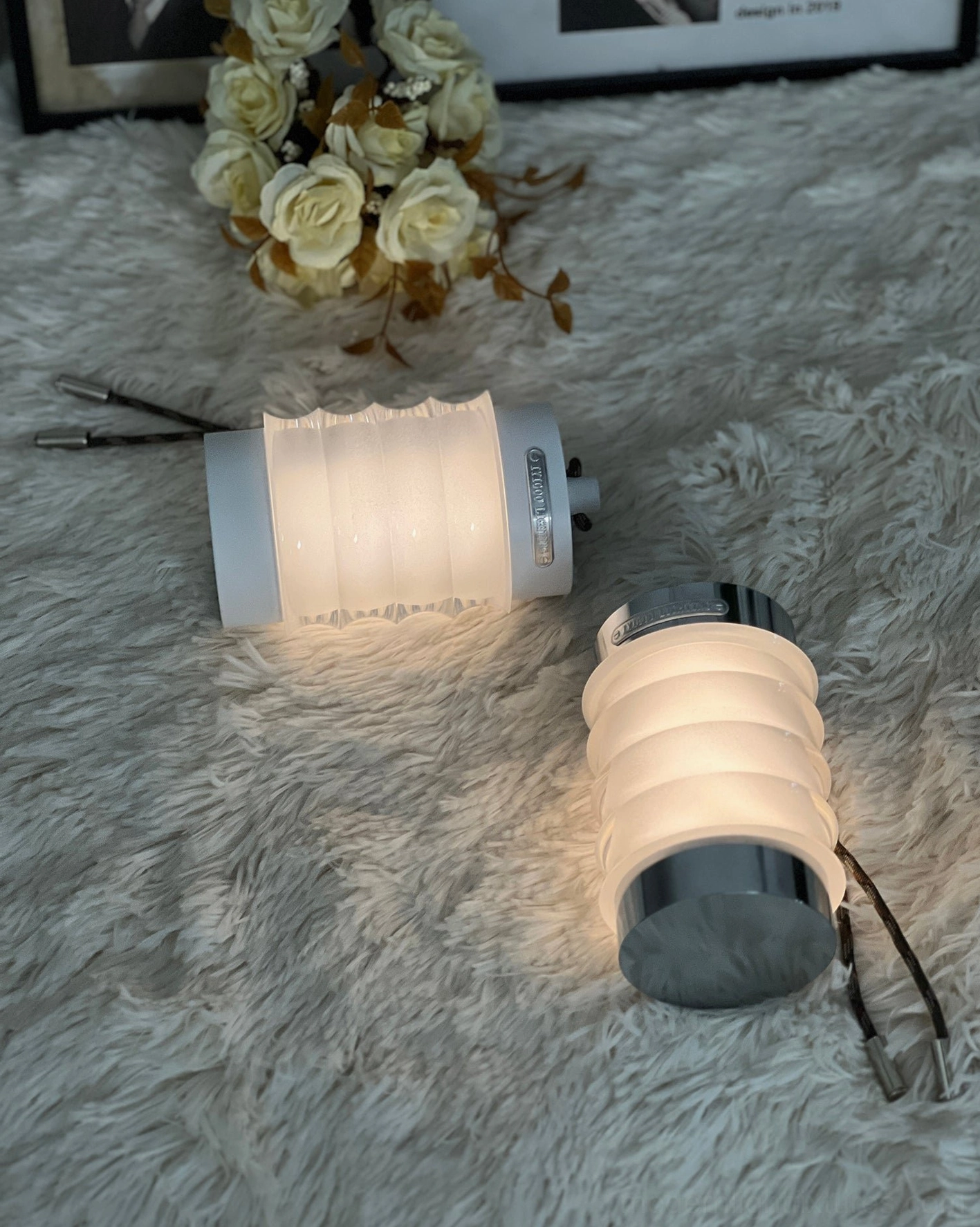 Pair of Balia portable LED table lamps in light gray and dark gray finishes, flat lay on fluffy fur rug with decorative roses