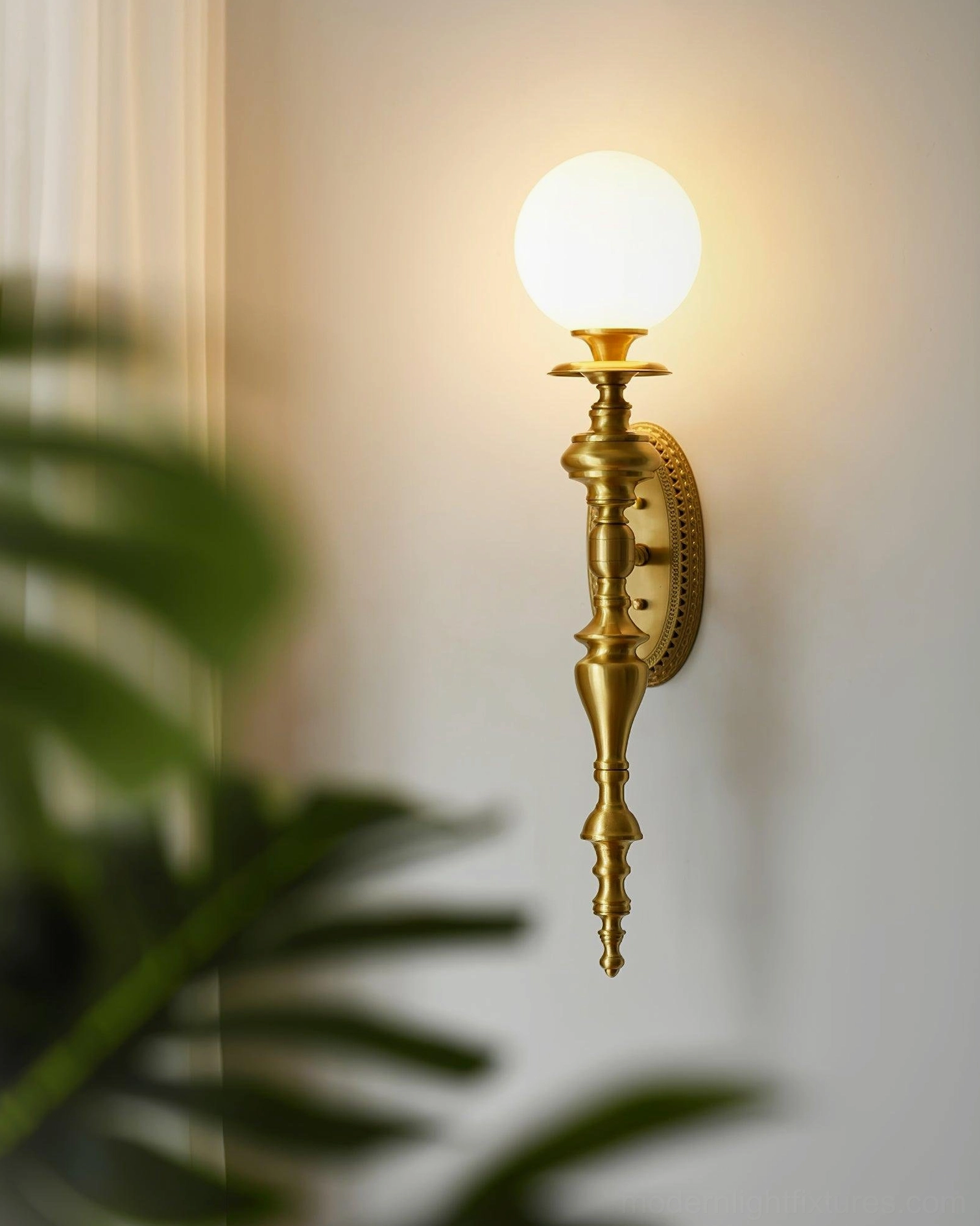 Brass wall sconce front view illuminated in living space with plant foreground and warm ambient glow