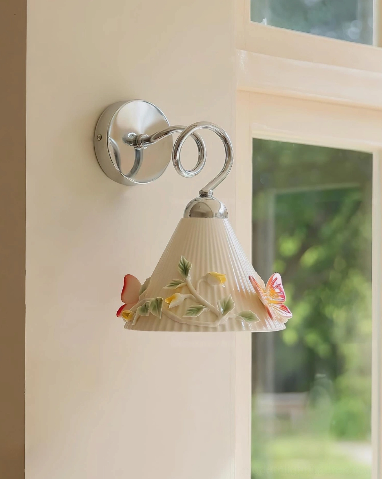 Ceramic butterfly wall sconce in bedroom interior with natural greenery visible, embossed butterflies and foliage on ribbed shade creating sculptural lighting