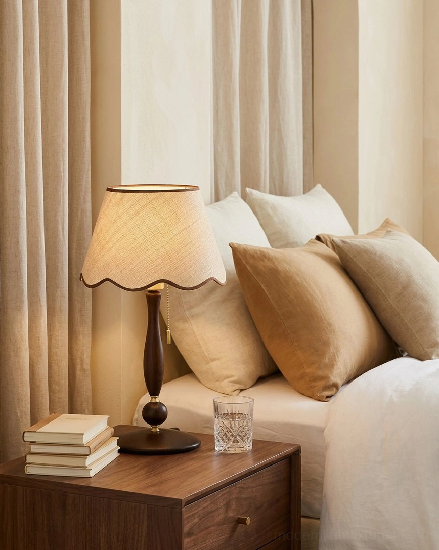 Bedside scalloped table lamp with cream linen shade on walnut wood base next to neutral tone bedding, stacked books and water glass