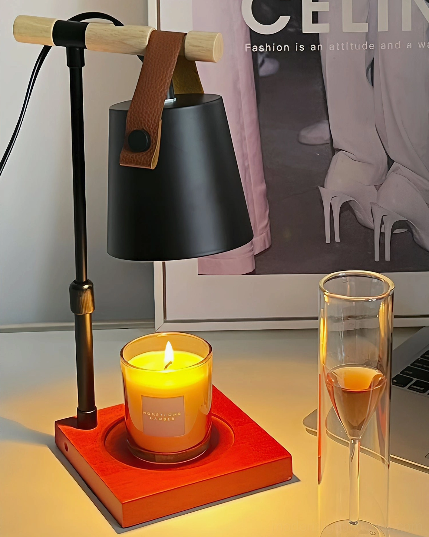 Black candle warmer lamp with red wood base, lit scented candle, wine glass and fashion poster on desk, evening ambiance