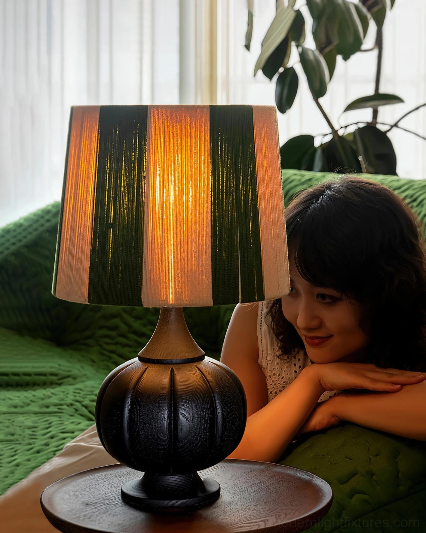 Black pumpkin base striped table lamp lit in cozy living room with green velvet sofa and woman resting