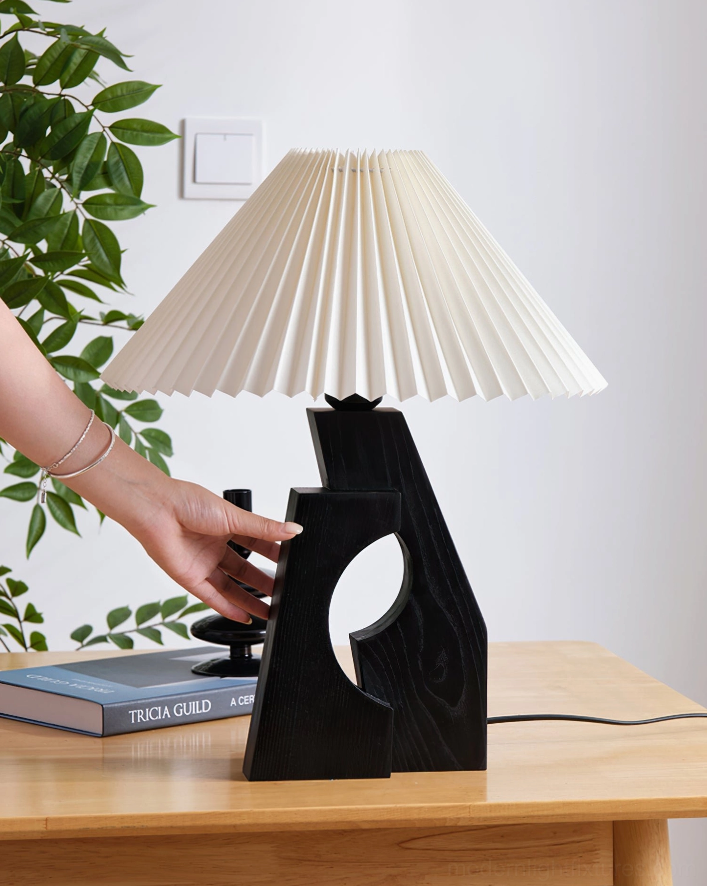 Black wood base pleated table lamp with hand interaction on home desk, surrounded by stacked books and green plant