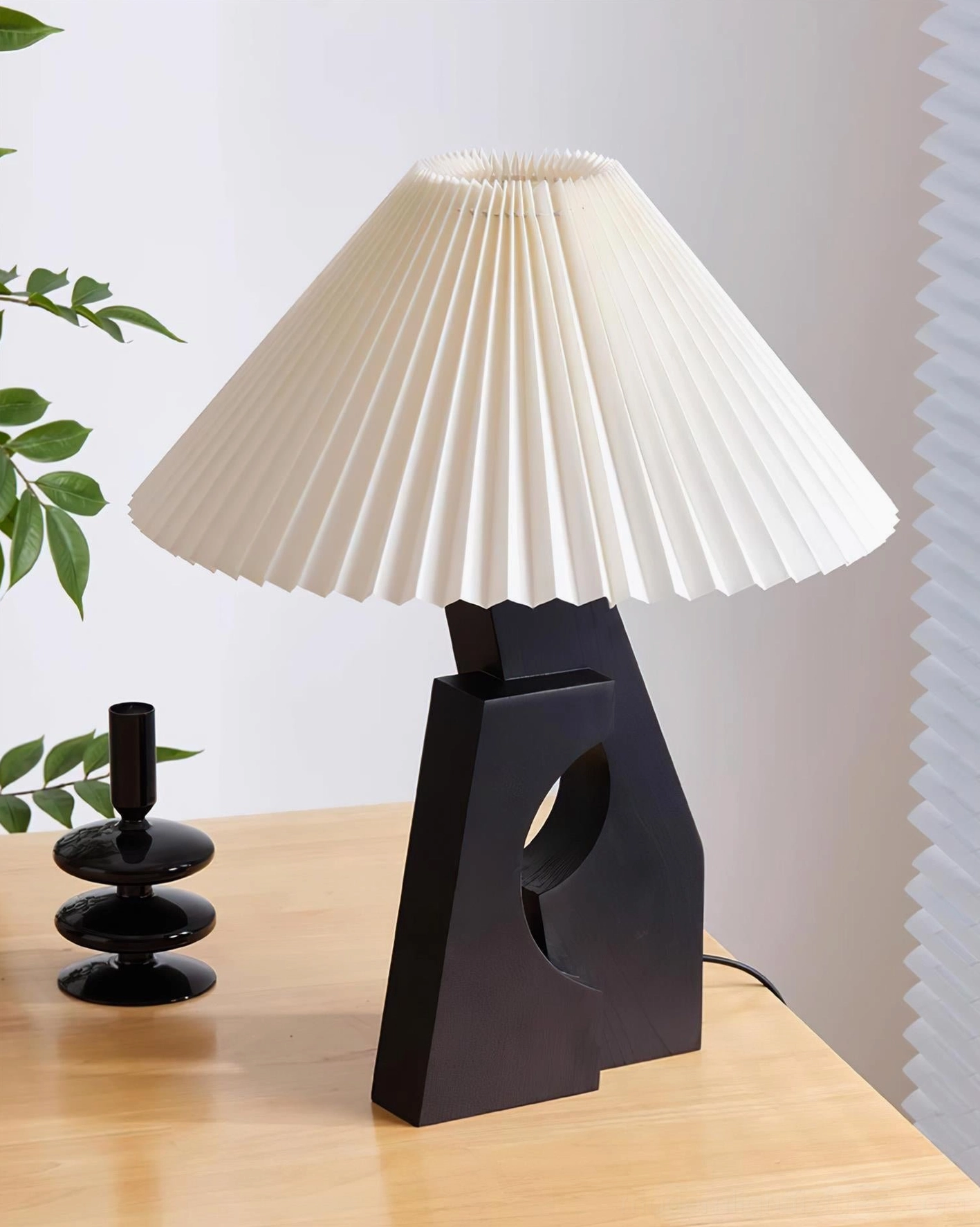 Unlit black hollow wood base pleated table lamp with white pleated shade, side view on wooden desk with candle decor
