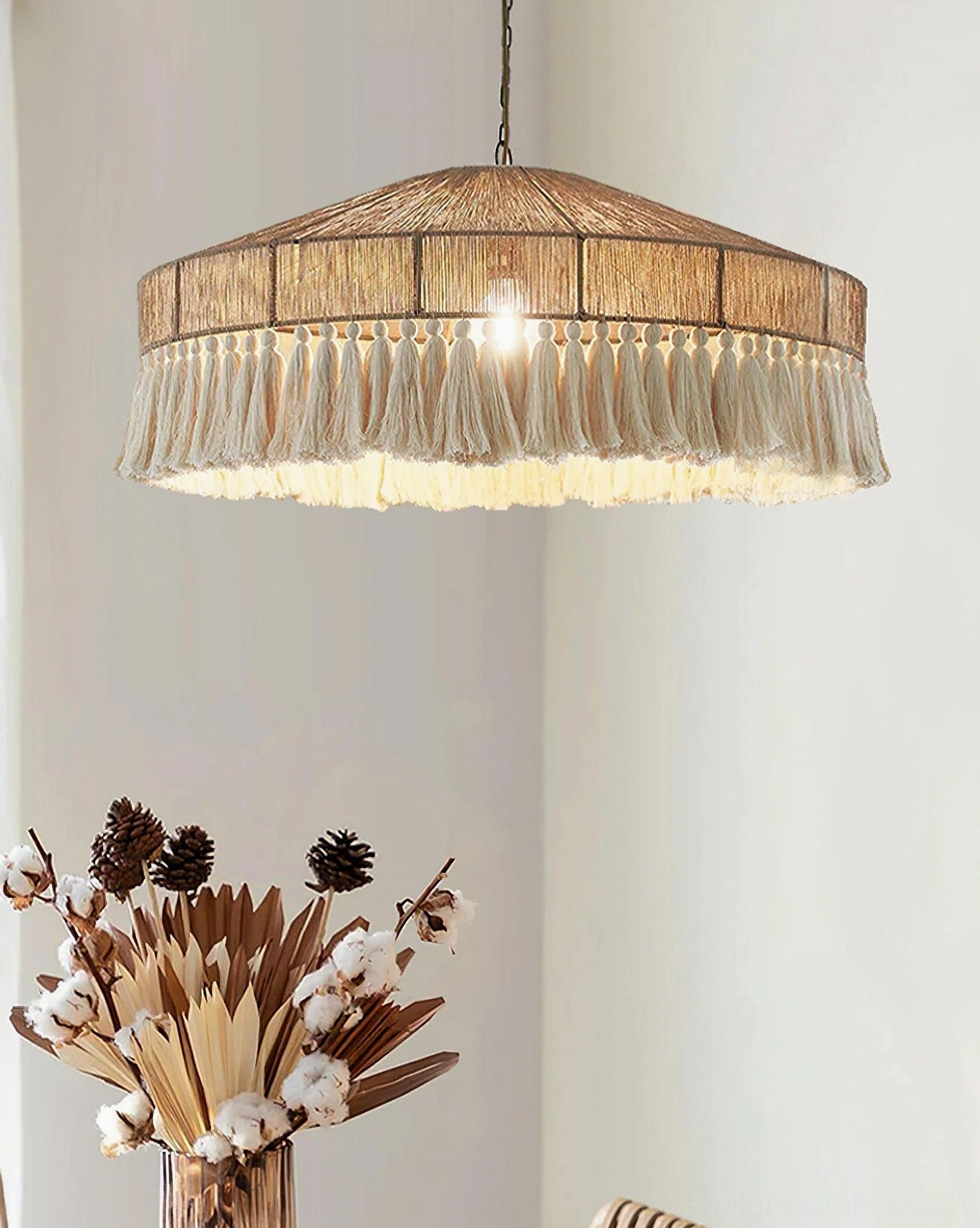 Bohemian tassel pendant light illuminated above dining area with dried flower arrangement, three-quarter view of lit warm glow and natural woven frame