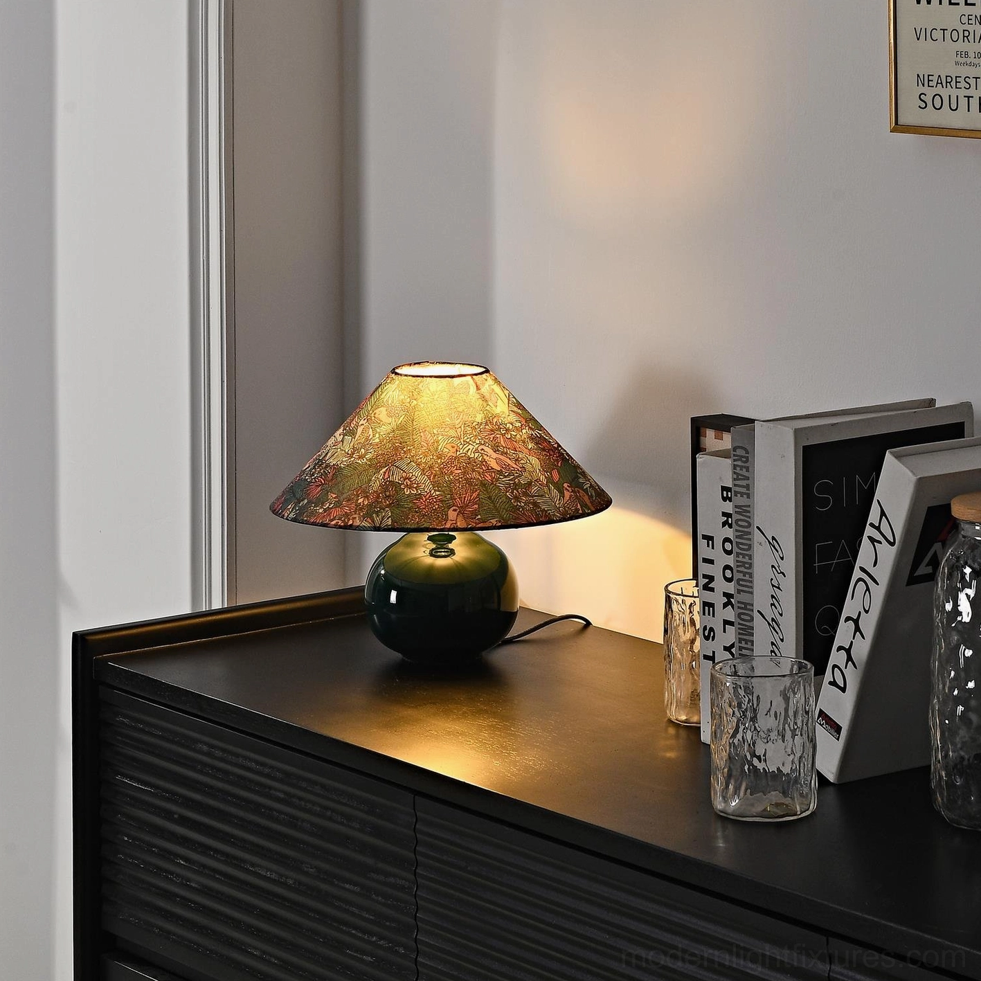 Botanical table lamp close-up showing visible power cord on dark wooden surface with warm illuminated shade and decorative styling