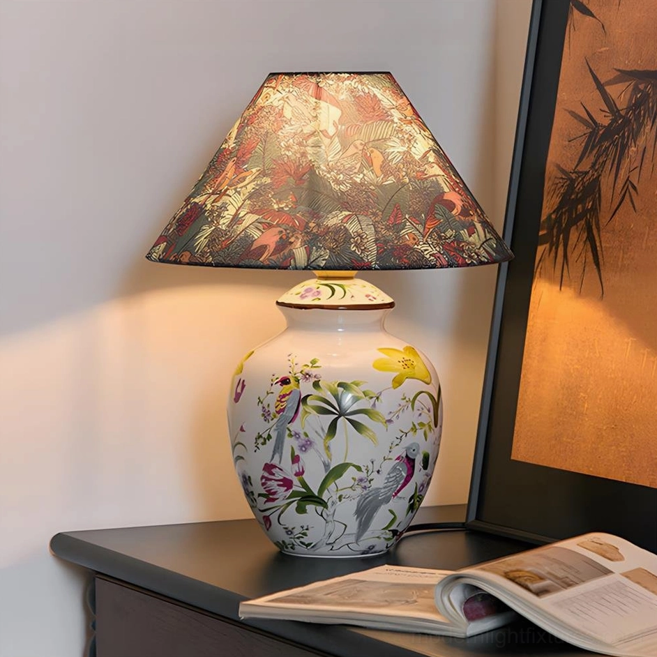 Table lamp with dark floral fabric shade and hand-painted ceramic base on dark desk with open book, front angle studio shot