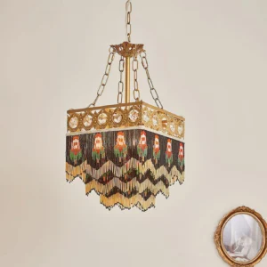 Brass square pendant lamp with handwoven beaded fringe and character motifs against decorative wall mirror