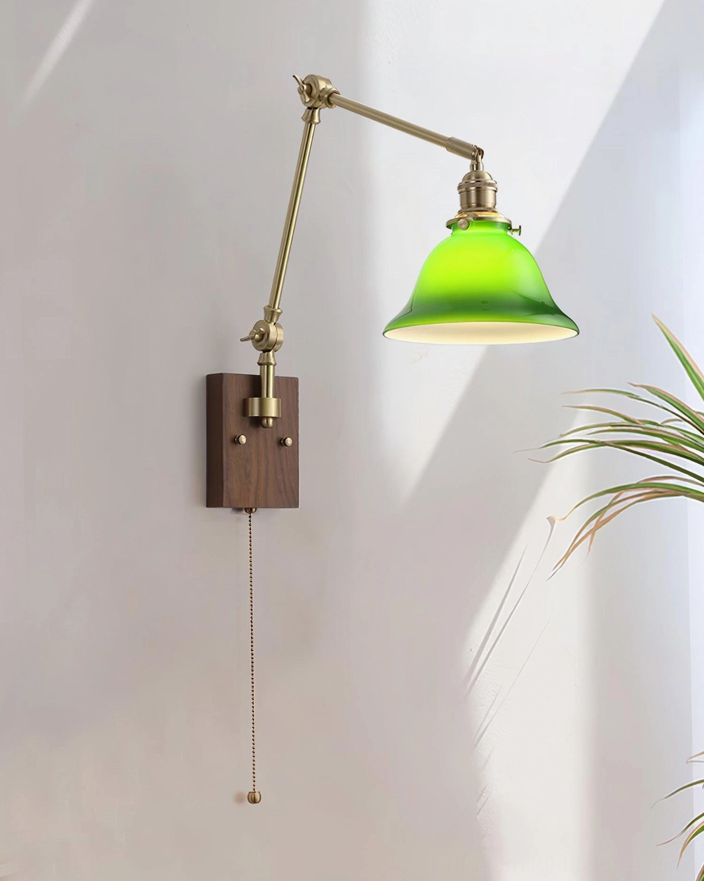 Side view of vintage brass swing arm lamp with green glass shade, walnut wall mount, and pull chain switch against sunlit wall with foliage
