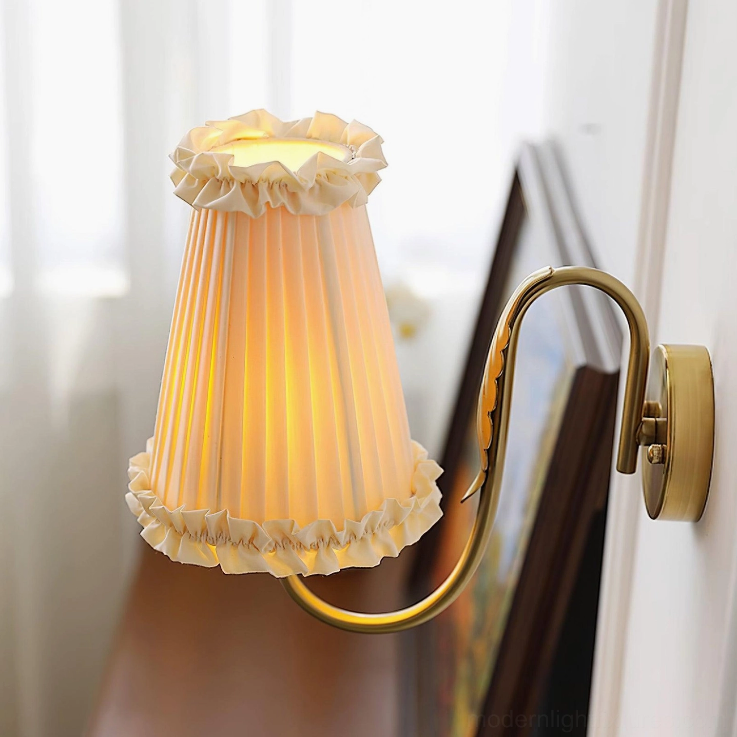Lit brass wall lamp with beige fabric shade above wooden furniture and artwork