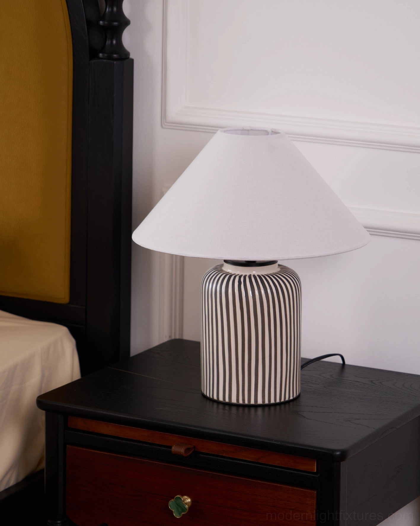 Three-quarter side view of black and white striped ceramic table lamp with tufted headboard backdrop