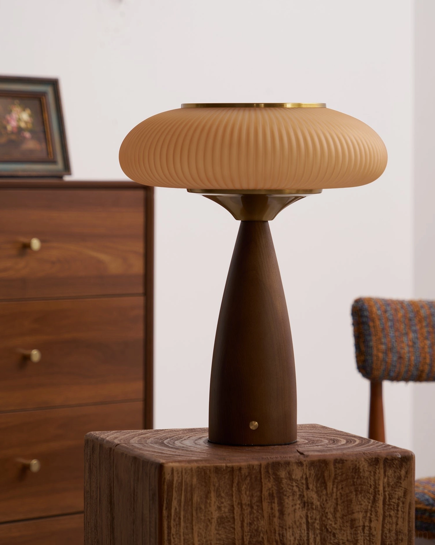 Brynn table lamp three-quarter left side view on wooden pedestal with walnut dresser and chair, warm ambient lighting