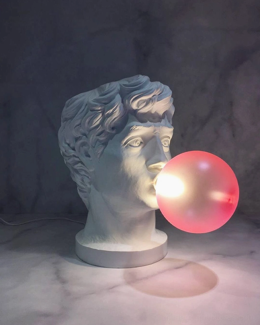 Bubblegum Boy resin lamp on marble tabletop with studio lighting and dark gray backdrop, showing warm pink glow
