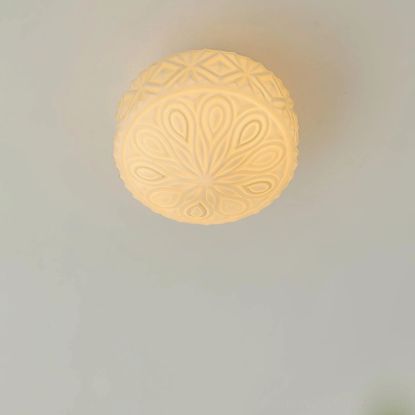 Lit vintage carved glass ceiling light fixture glowing warm yellow, front ceiling view on plain white indoor wall
