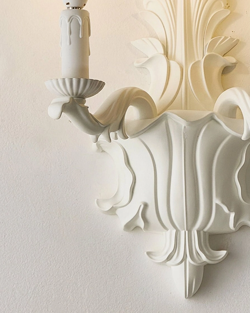 Matte white carved wall sconce backlighting acanthus-leaf detail with warm upward-pointing glow