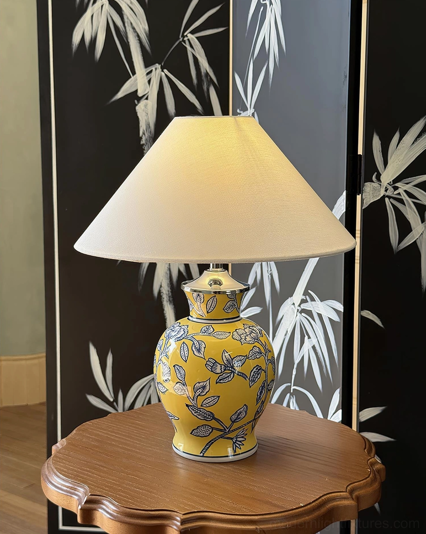 Yellow ceramic table lamp with blue flower pattern and fabric shade, placed on carved wooden round side table