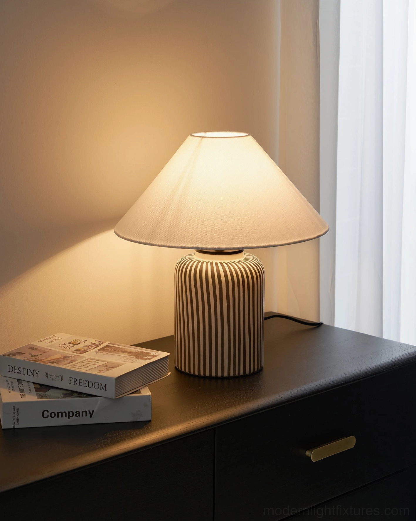 Hand-painted vertical striped ceramic lamp on wooden table with stacked books and sheer curtain background
