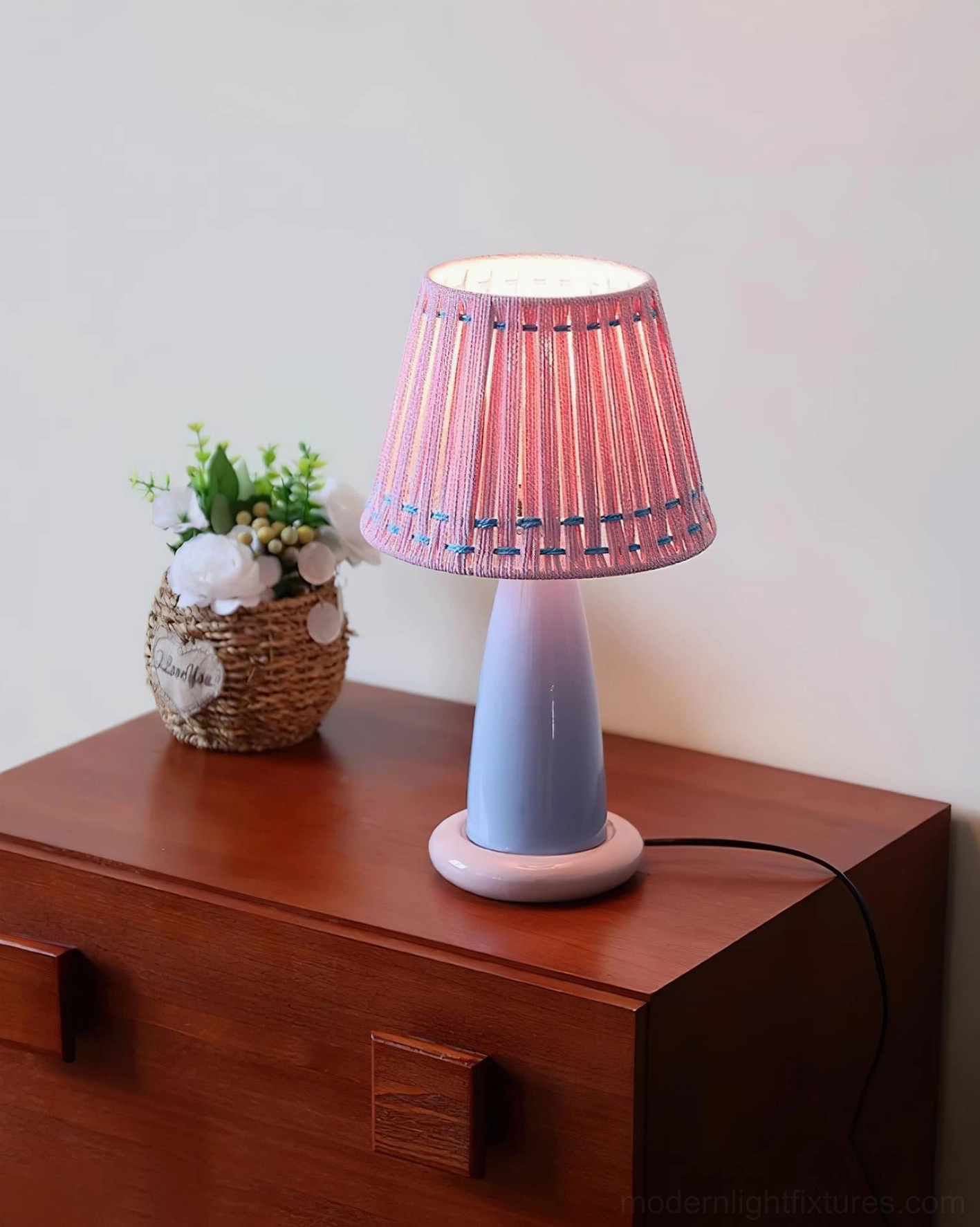 Pink woven shade ceramic table lamp illuminated on wooden dresser with flower basket, cozy bedroom lighting atmosphere