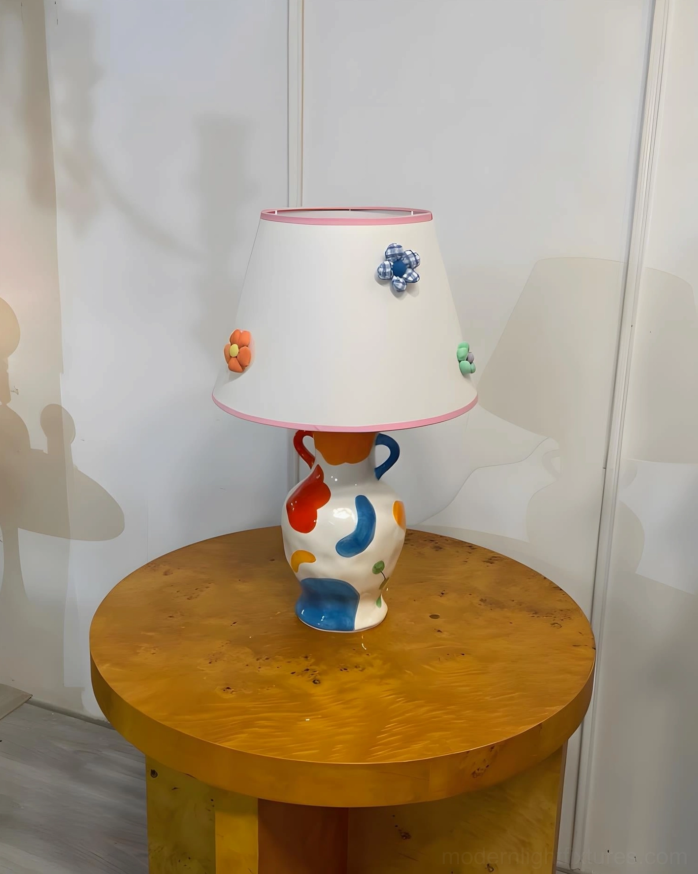 Ceramic table lamp frontal three-quarter perspective on wooden round table, displaying colorful painted base and flower-adorned white fabric shade