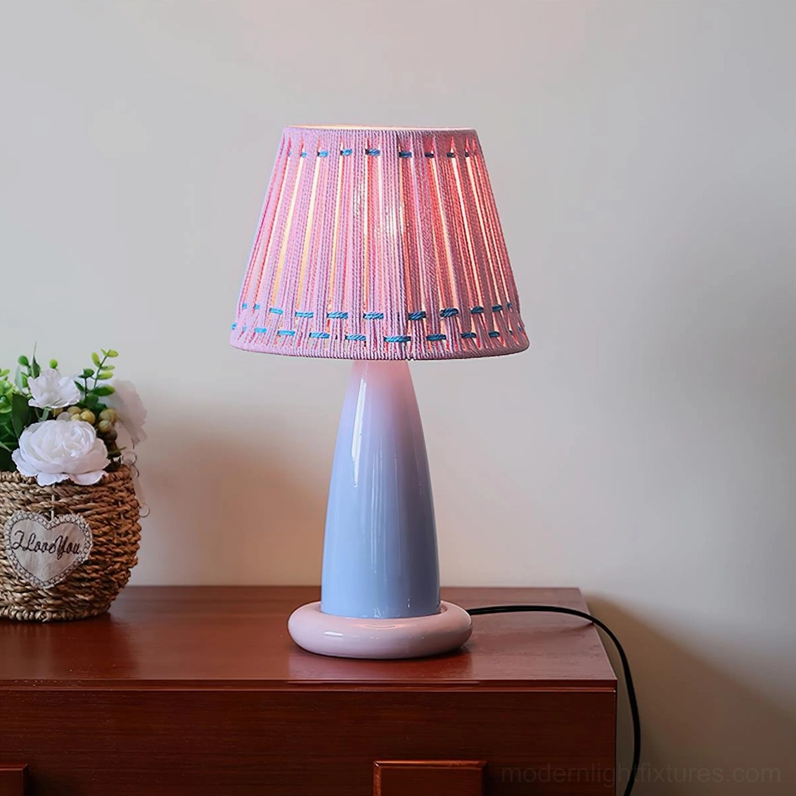 Pink woven shade ceramic table lamp displayed on wooden dresser with decorative accents, soft pastel lighting fixture