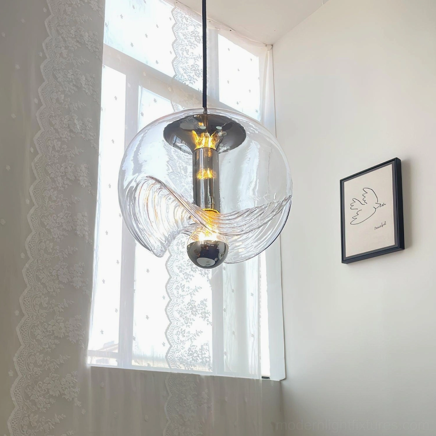Clear glass wave pendant light by bright window with lace curtains, close-up showing contemporary minimalist design and polished metal components