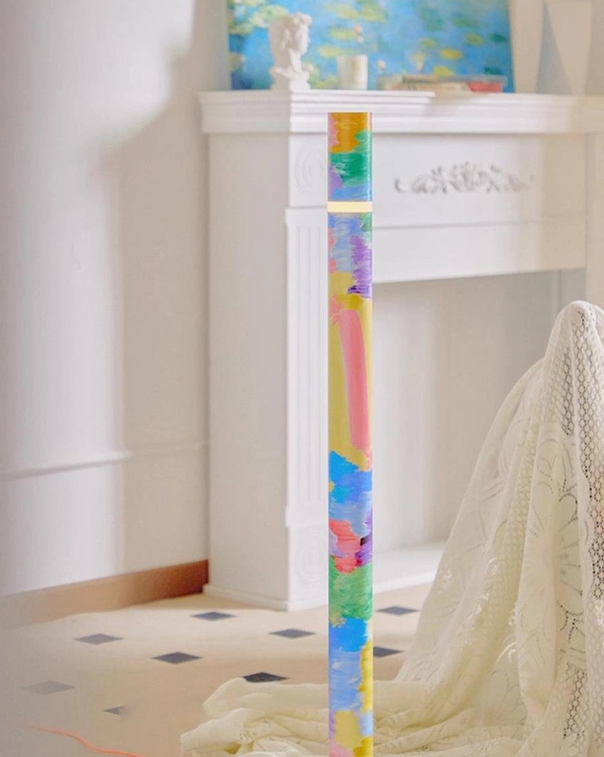 Colorbeam painted floor lamp close-up detail in living room near fireplace, showing vibrant hand-painted cylinder with lace throw on nearby chair