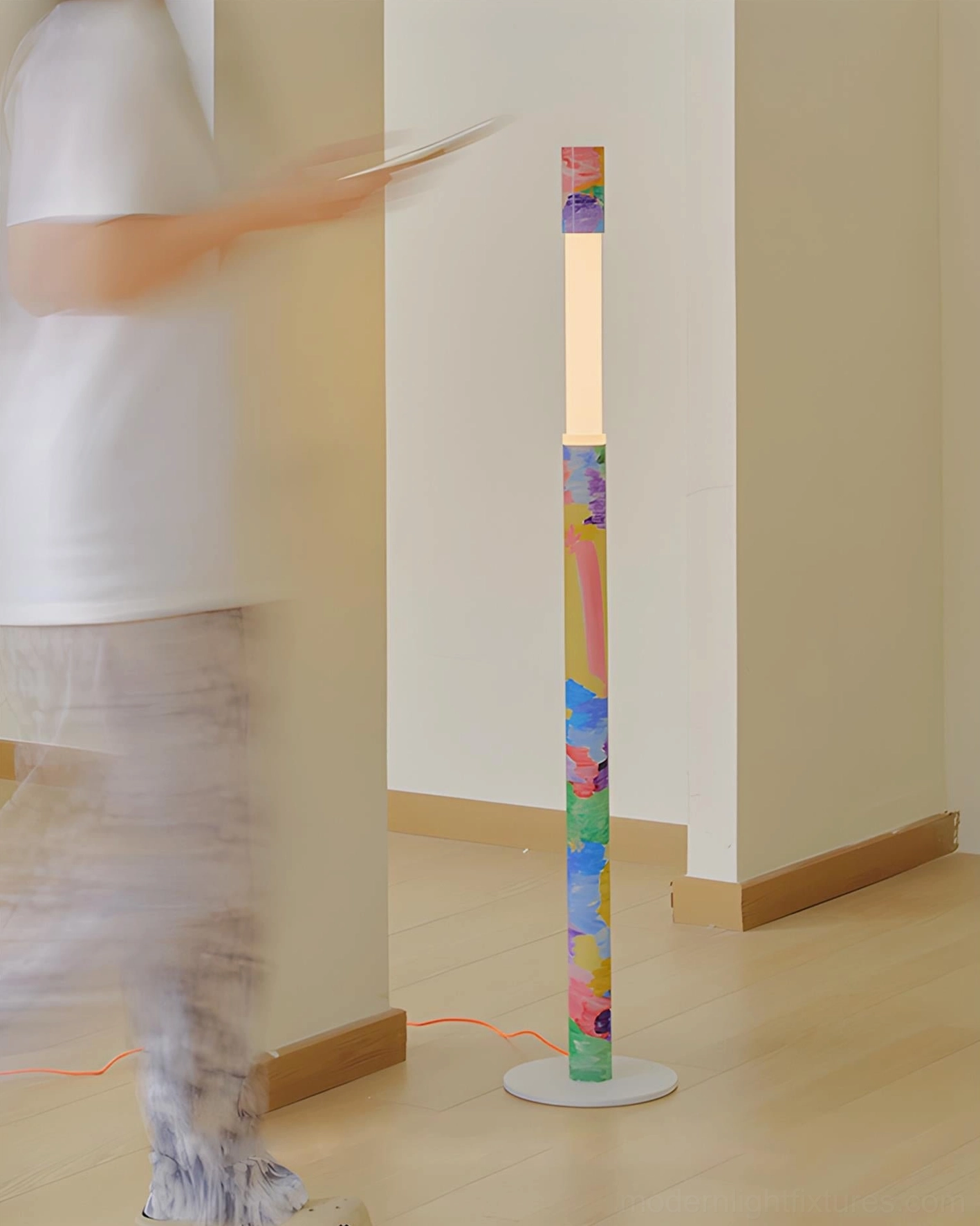 Colorbeam floor lamp in minimalist apartment hallway side view with blurred figure walking past, showcasing tall sculptural presence and red power cord