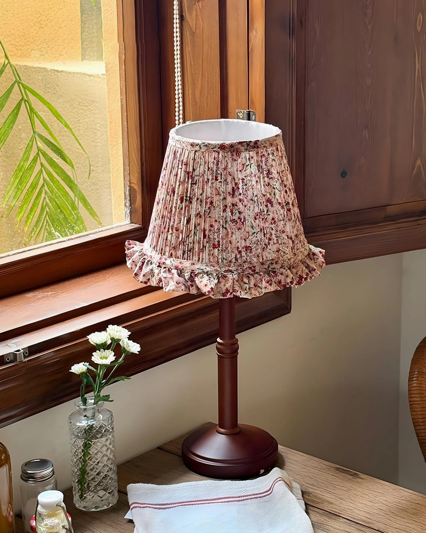 Cottage table lamp placed on dining table by vintage window with flower vase and linen napkin, side view