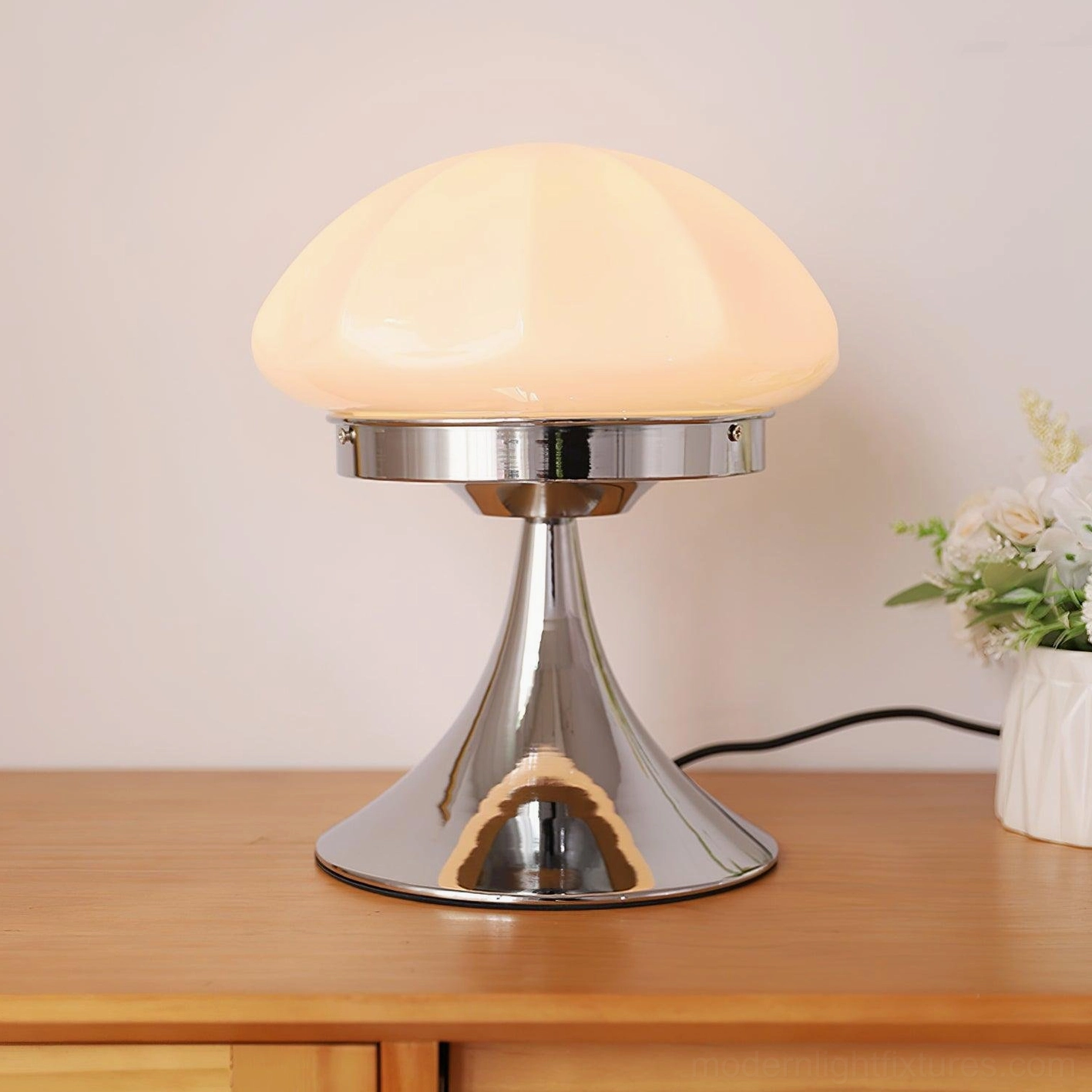 Illuminated Dario mushroom table lamp with beige glass shade glowing warmly on wooden nightstand with flowers