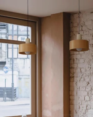 Two wooden Dovira drum pendant lights suspended in residential apartment corner with exposed brick, warm neutral decor, and natural window light