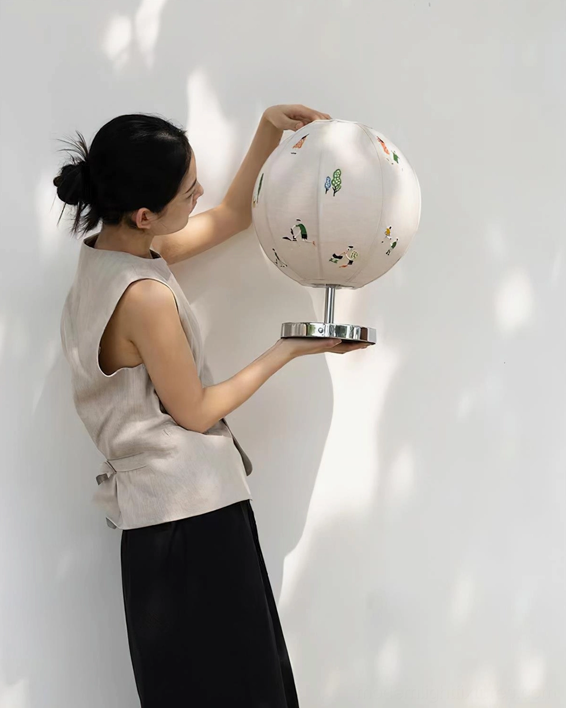 Narrative embroidered table lamp in profile against sunlit wall, woman holding fixture displaying fabric embroidery detail