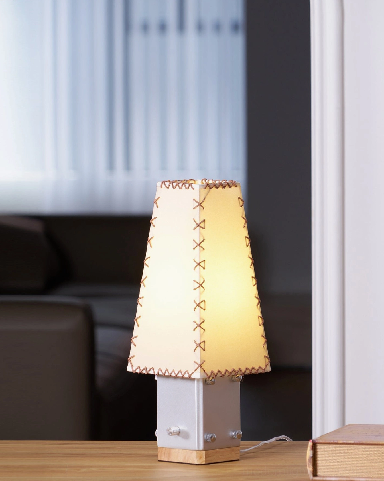 Estela table lamp glowing warmly in living room corner with dark sofa and blinds