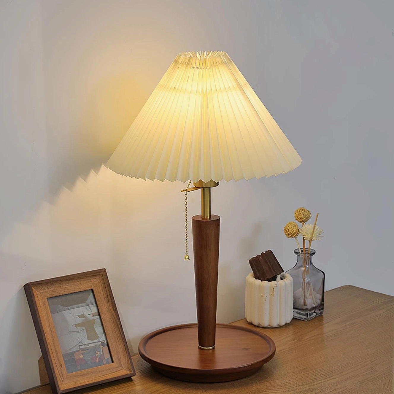 Pleated fabric table lamp with brass-finished hardware creating warm yellow glow on side table with framed photo