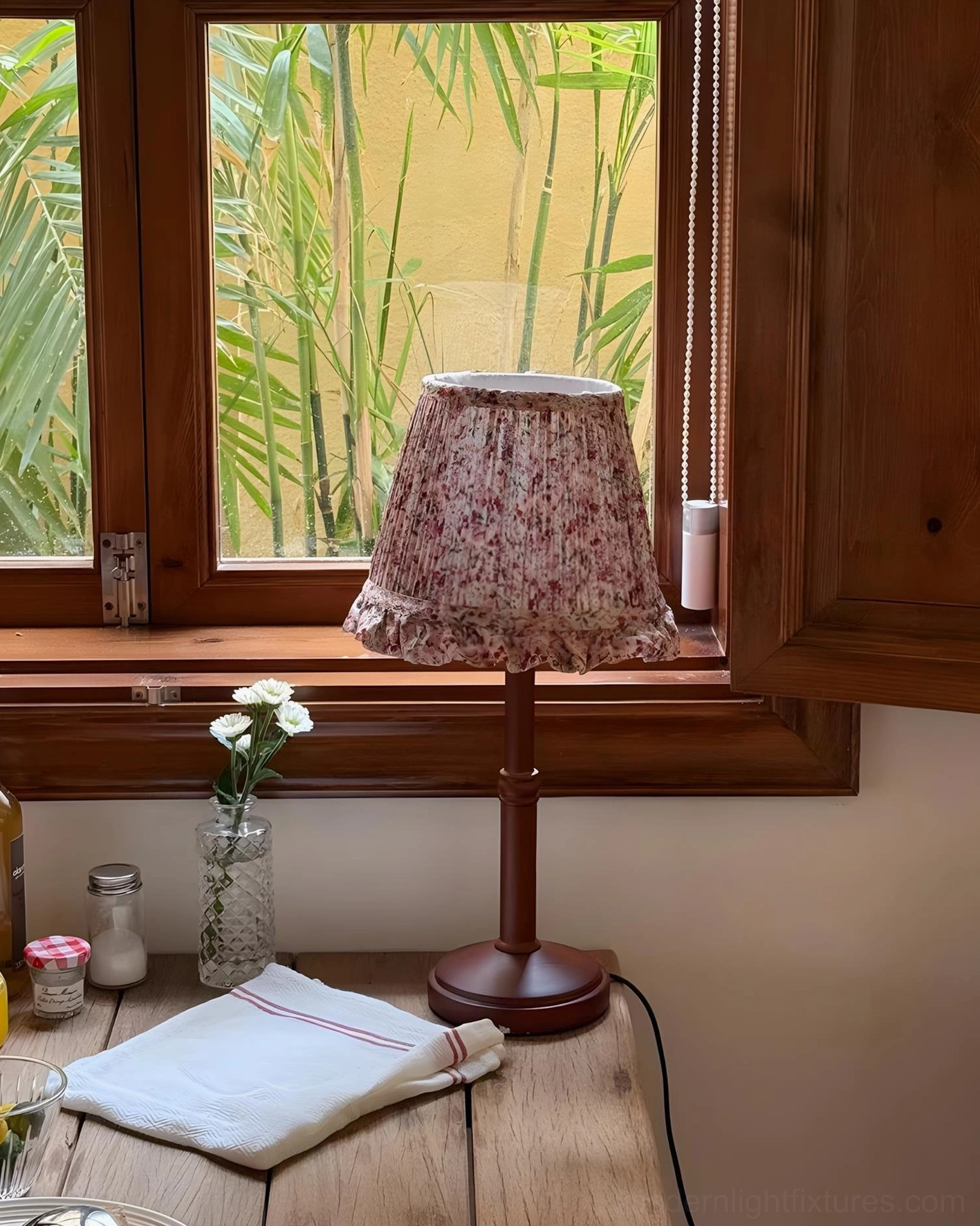 Floral pleated table lamp on wooden table by brown window with glass flower vase and linen