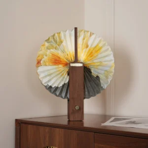 Folding paper table lamp with pleated watercolor shade displayed on bedroom wooden nightstand with papers and minimalist decor